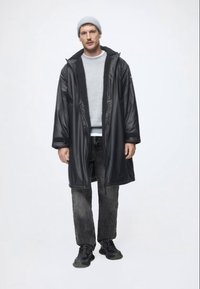 Cagulă neagră, impermeabilă, cu guler din blană, purtată peste un pulover gri și pantaloni texturați închisi. Modelul poartă ghete atletice negre și o caciulă gri.