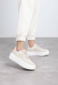 Zapatillas beige con cordones, con un empeine texturizado, suela blanca plana y acentos naranjas cerca del talón, sobre un fondo neutral.