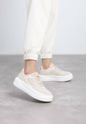 Zapatillas beige con cordones, con un empeine texturizado, suela blanca plana y acentos naranjas cerca del talón, sobre un fondo neutral.
