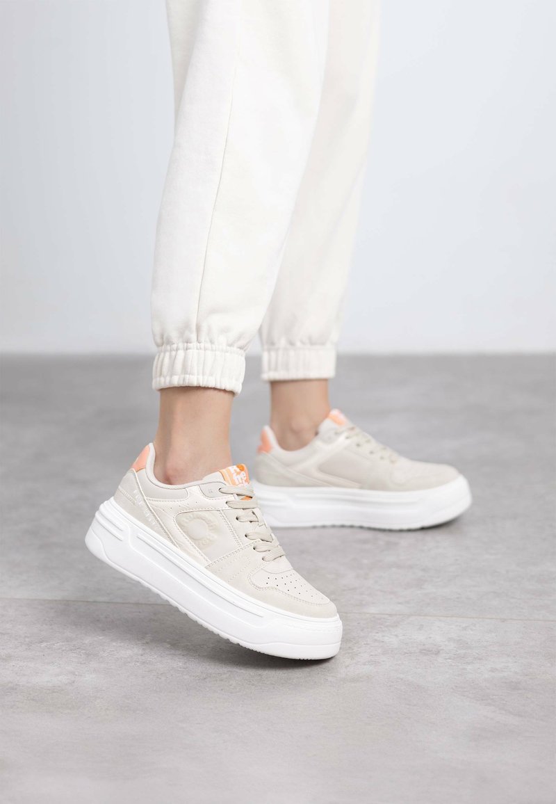 Zapatillas beige con cordones, con un empeine texturizado, suela blanca plana y acentos naranjas cerca del talón, sobre un fondo neutral.