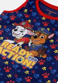 Blauw tanktop met cartoonhonden in rode en blauwe uniformen. Tekst "KLAAR VOOR ACTIE!" omringd door kleurrijke pootafdrukken. Rode kraag.