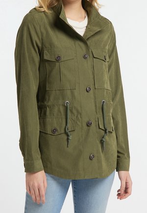 Femme portant une veste boutonnée vert olive avec poches à rabat et cordons réglables à la taille, assortie à un jean bleu clair.