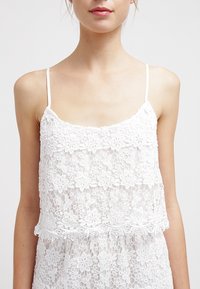 Bílá krajková košilka s květinovým vzorem, jemnými ramínky a vrstvenou texturou. Měkký, lehký materiál, ideální pro vrstvení nebo teplé počasí.