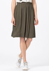 Zelená olivová sukně do délky kolen s volnými plisováními, elastickým pasem a hladkou texturou látky. V kombinaci s bílými teniskami.