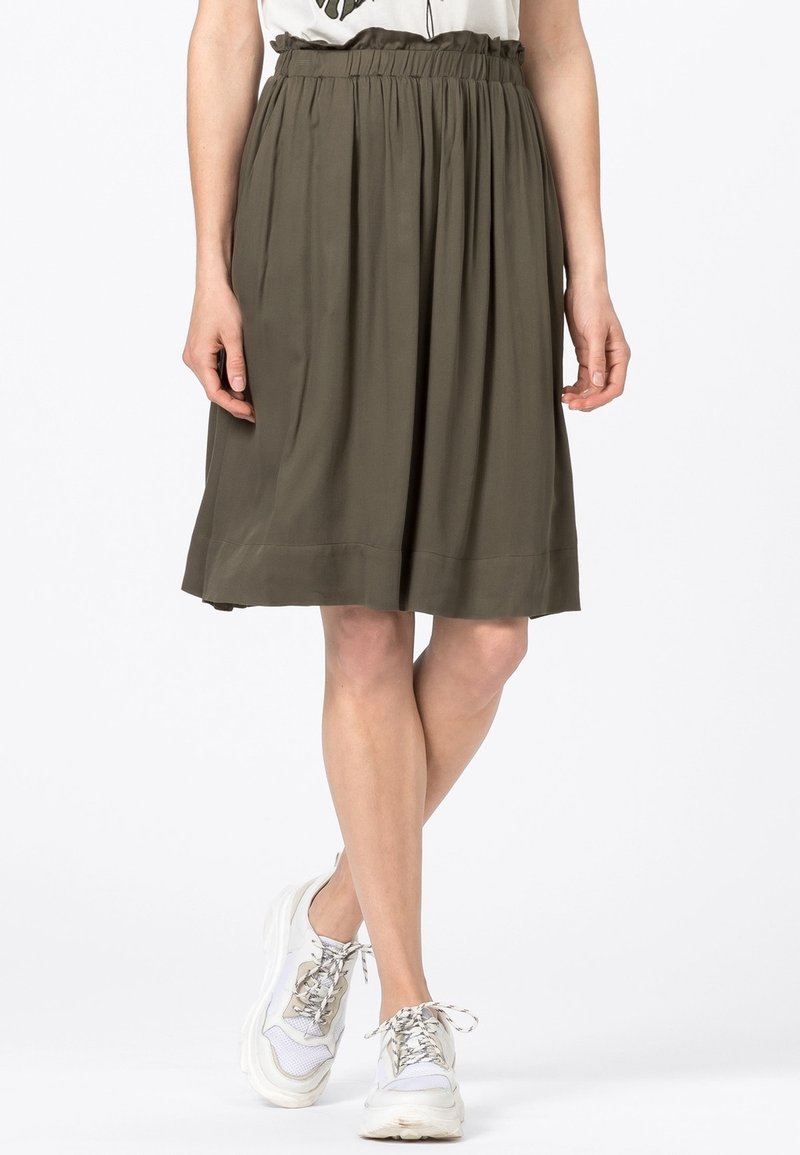 Zelená olivová sukně do délky kolen s volnými plisováními, elastickým pasem a hladkou texturou látky. V kombinaci s bílými teniskami.
