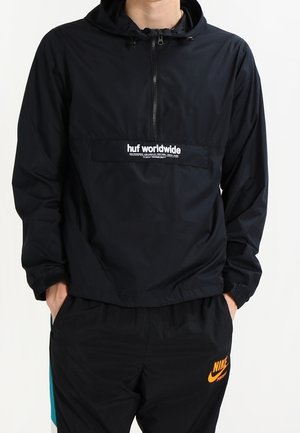 Mężczyzna w czarnej kurtce z kapturem na półzamek z logo "huf worldwide" oraz czarnych spodniach Nike z pomarańczowym logo Swoosh.