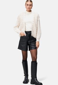 Crème cardigan met textuurfranje, witte turtleneck, zwarte leren shorts en kniehoge zwarte laarzen, tegen een witte achtergrond.