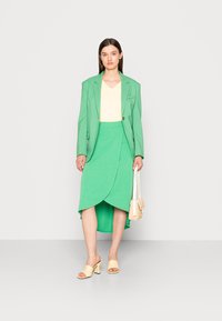 Blazer verde sobre uma blusa amarela, combinado com uma saia envolvente verde e sandálias de salto amarelas. Segurando uma pequena bolsa de palha.