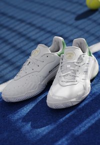 Coppia di scarpe da tennis bianche Adidas con dettagli verdi su un campo da tennis blu vicino a una palla e a una linea bianca.