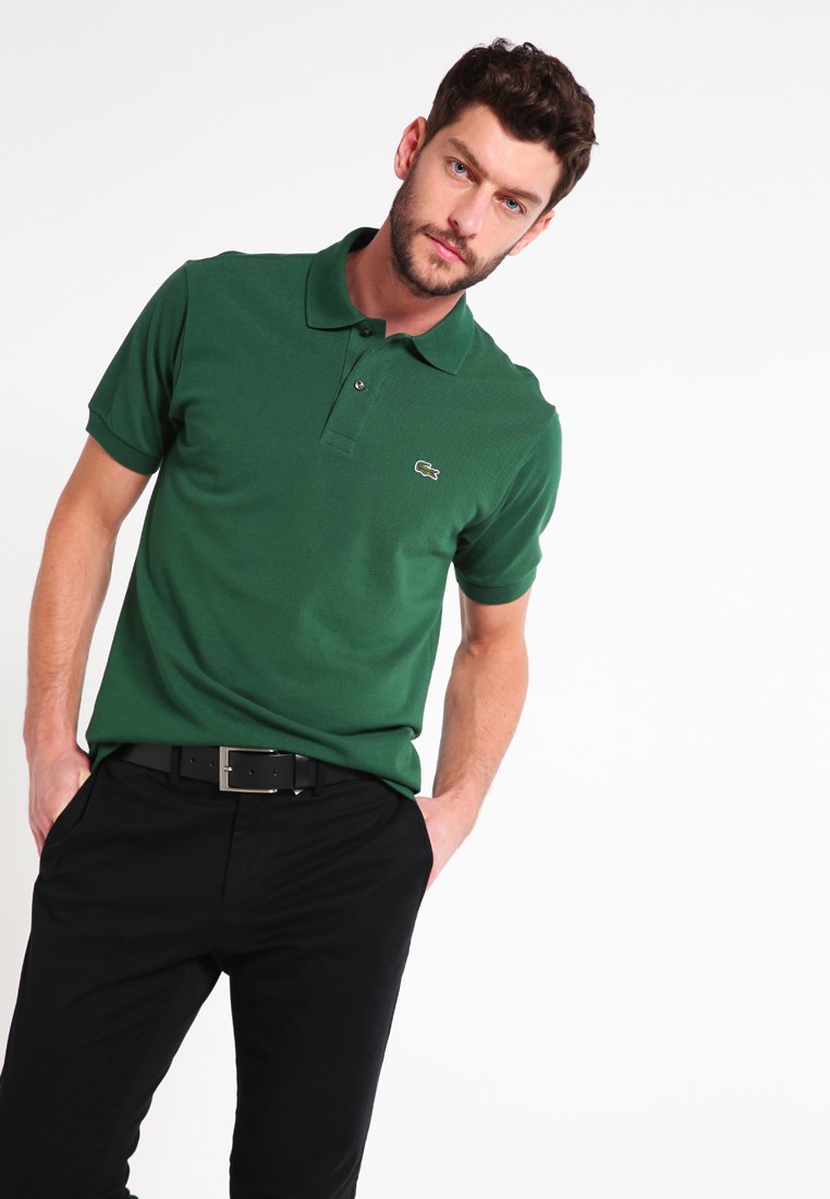 Camisa polo de algodón verde con un cuello clásico, mangas cortas y un pequeño logo bordado en el lado izquierdo del pecho. Combinada con pantalones negros.