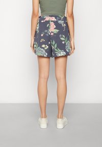 Žena oblečená v námořnicky modrých květovaných šortkách, olivově zeleném zkráceném topu a bílých platformových teniskách, stojící zády k bílé pozadí.