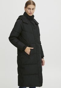 Svart lang dunjakke med høy krage, glidelåslommer og quiltet design med en jevn tekstur og kroppsnær silhuett.