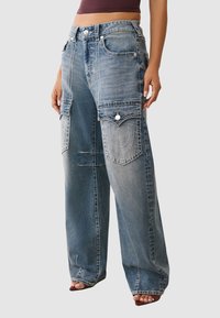 Jasnoniebieskie luźne dżinsy z wieloma naszywanymi kieszeniami, kontrastowymi szwami oraz wysokim stanem. Teksturowana tkanina o zużytym wyglądzie.