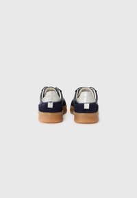 Mørkeblå ruskinds sneakers med en gummisål, der har en sølvfarvet hælflap, kontrastsying og et struktureret design på bagsiden.