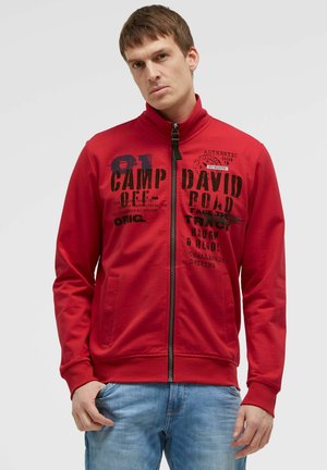 Man with short brown hair wearing a red zip-up jacket with black text and blue jeans against a plain background.