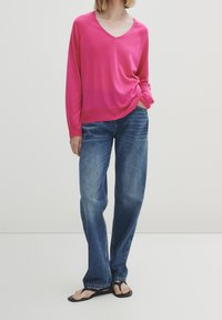 Pull fuchsia à col en V associé à un jean en denim bleu ample et des tongs noires ; présente une coupe décontractée et des poignets côtelés aux manches.