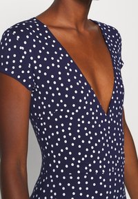 Mörkhyad person klädd i en navyblå klänning med vita prickar och djup V-ringning, sedd från axlarna till midjan.