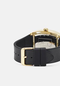 Must mustriba mustran pärga kullast metallist klambriga, millel on tekstureeritud aluspind ja ristküliku kujuline kellanägu koos nähtava brändi logoga ja detailidega.