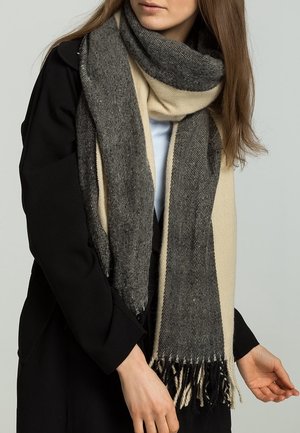 Femme portant une longue écharpe épaisse beige et gris foncé avec des franges sur un manteau noir, les mains visibles au niveau de la taille.