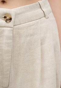 Κοντινό πλάνο μπεζ παντελονιού από λινό με ψηλή μέση, κλείσιμο με κουμπί και θηλιές για ζώνη, που αναδεικνύει την υφή του υφάσματος και τους φυσικούς τόνους.