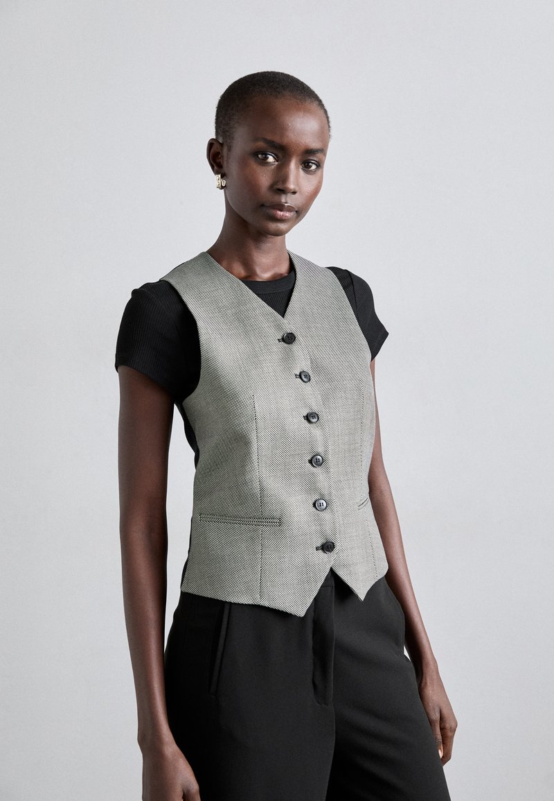 Pelēks houndstooth veste ar V formas izgriezumu, sešām melnām pogām un divām priekšējām kabatām, uzvilkta virs melnas īsām piedurknēm tērpa.