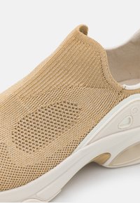 Bequemer Sportschuh mit einem strukturierten beigen Strick-Obermaterial, das ein Netzpanel sowie eine weiße und goldene Gummisohle mit Logo-Details aufweist.