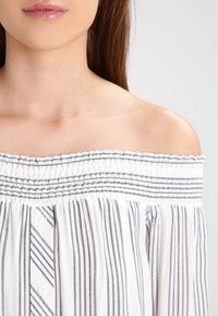 Off-shoulder blus gjord av lättviktsmaterial, med vertikala svart-vita ränder, smockad halsringning och knapprad.