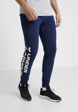 Mann trägt marineblaue Under Armour Jogginghose mit Logo am linken Bein und schwarze Sportschuhe mit weißen Sohlen.