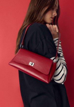 Frau in schwarzem und gestreiftem Oberteil, die eine rote Leder-Umhängetasche mit silberner Schnalle vor rotem Hintergrund hält.