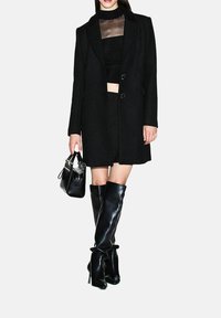 Mujer con un abrigo largo negro, blusa negra transparente, minifalda, botas negras brillantes hasta la rodilla y sosteniendo un bolso negro sobre un fondo blanco.