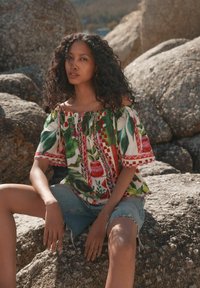 Blusa sin hombros en blanco con hojas verdes, acentos rojos y patrones florales; combinada con shorts de mezclilla desgastados.