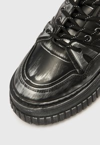 Melnas sporta kurpes ar spīdīgu virsmu, teksturētām detaļām un smalku auklu izstrādājumu. Raksturīgs skrāpējumu raksts pirkstu zonā. Gumijas zole.