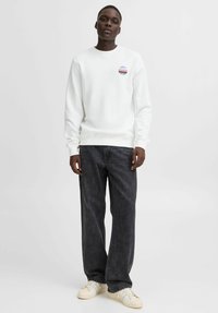 Witte sweatshirt met een ronde halslijn en een kleine, kleurrijke grafische afbeelding; gecombineerd met donkere, losse spijkerbroek en lichte sneakers.