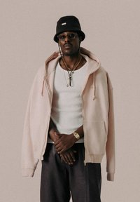 Sweat à capuche rose oversize en tissu doux, porté par-dessus un débardeur blanc, associé à un pantalon sombre et un chapeau seau noir.