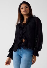 Czarna plisowana bluzka z falbanianym kołnierzem i zamknięciem z przodu, długimi rękawami oraz wycięciem. Noszona z jasnoniebieskimi dżinsami.
