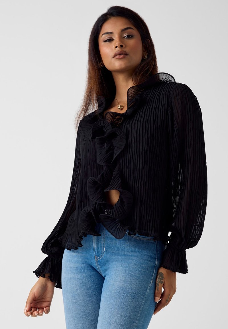 Czarna plisowana bluzka z falbanianym kołnierzem i zamknięciem z przodu, długimi rękawami oraz wycięciem. Noszona z jasnoniebieskimi dżinsami.