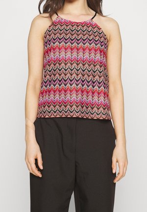 Femme portant un haut sans manches avec un motif zigzag multicolore et un pantalon noir ample, debout devant un fond uni.