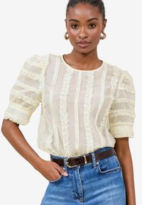 Cream blouse featuring vertical stripes and ruffled accents, short puff sleeves, sheer texture, paired with dark denim and a brown belt.