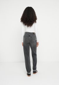 Femme aux cheveux bouclés portant un haut blanc à manches longues, un jean noir délavé taille haute et des baskets noires, debout face à un mur blanc.