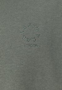 Grøn strikket stof med et cirkulært broderet design, hvor ordene "SAINT L" og "ONDON" er integreret, hvilket skaber en tekstureret visuel interesse.