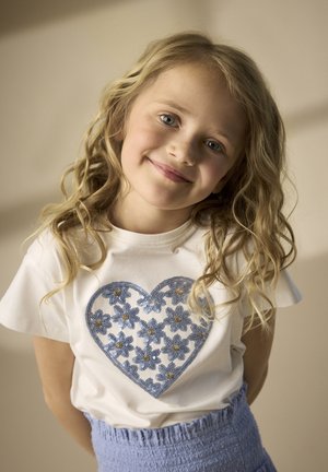 Hvid T-shirt med korte ærmer og et blåt hjerte-design, der har blomsterformede pailletter og gyldne accenter. Bær med et lyseblåt tekstureret skørt.