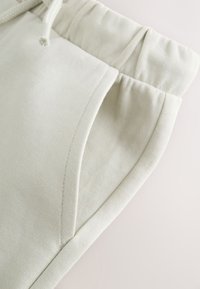 Gros plan de la zone de poche d'un pantalon de survêtement beige clair avec taille élastique et extrémité de cordon visible.