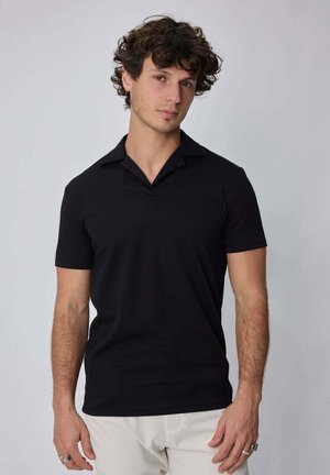 Jeune homme aux cheveux bouclés portant une chemise noire à manches courtes avec col et un pantalon de couleur claire, debout devant un fond uni.