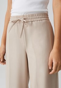Pantalons beige à jambes larges avec une taille élastique et un cordon de serrage. Le tissu a une texture lisse avec des rayures verticales subtiles. Accompagné d'un haut ajusté.