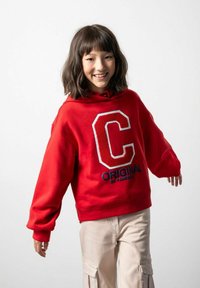 Czerwony sweatshirt z kapturem z dużym białym logo "C" oraz napisem "ORIGINAL". Wykonany z bawełny, luźny krój, z ribbowymi mankietami. Zestawiony z beżowymi spodniami cargo.