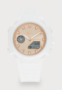 Montre Lacoste avec un bracelet en silicone blanc, un cadran beige rond, des accents dorés, affichage analogique et numérique, et un détail de logo à 12 heures.
