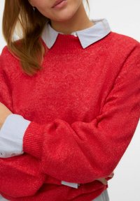 Jersey rojo de punto con cuello redondo y puños acanalados, superpuesto sobre una camisa de color azul claro con cuello. Textura suave con un ligero brillo.