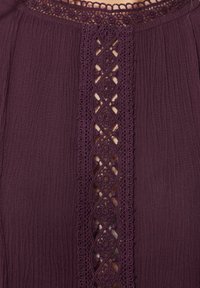 Blusa viola con un tessuto testurizzato, caratterizzata da pannelli di pizzo intricati lungo il centro. Il colletto presenta delicate rifiniture in pizzo.