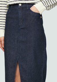 Jeansrock mit hohen Schlitz, dunkelblauer Farbe, Fronttasche und Metallknopfdetail. Der Stoff ist glatt und robust.