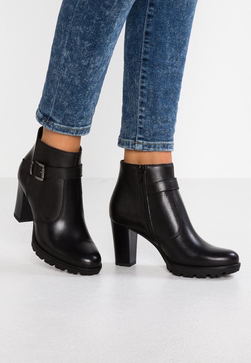 Botas de tobillo de cuero negro con un tacón grueso, que cuentan con un zipper lateral, una correa decorativa con una hebilla y una suela texturizada.